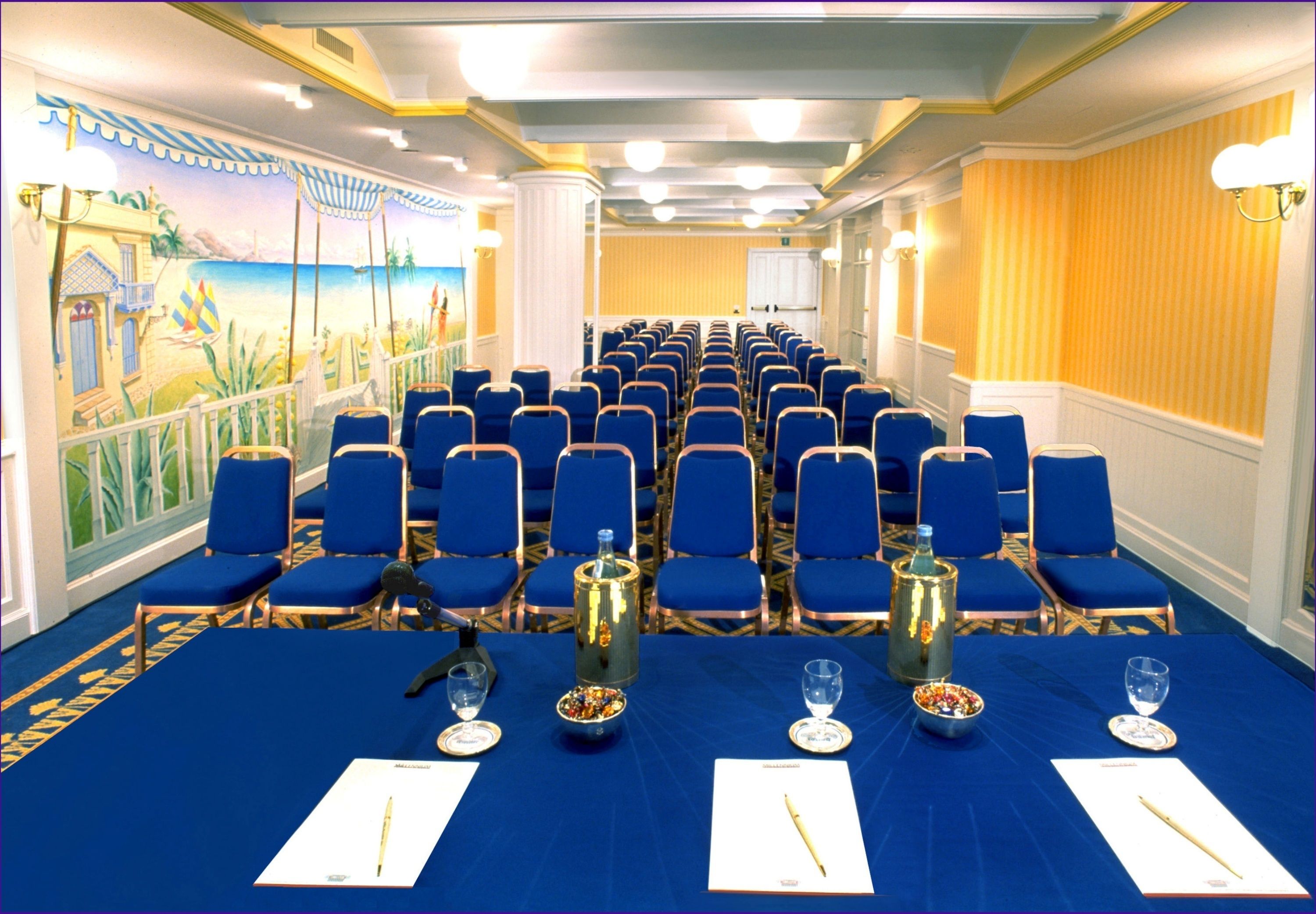 Ein Konferenzraum mit Reihen blauer Stühle, einem langen Tisch mit Namensschildern und einem Wandgemälde mit Strandmotiv.