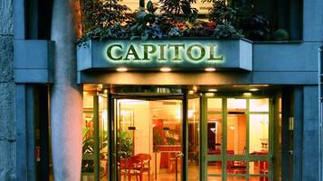 Een hotelentree met een glazen deur, een bord met de tekst 'CAPITOL' en een bloembak met verschillende planten boven de ingang.