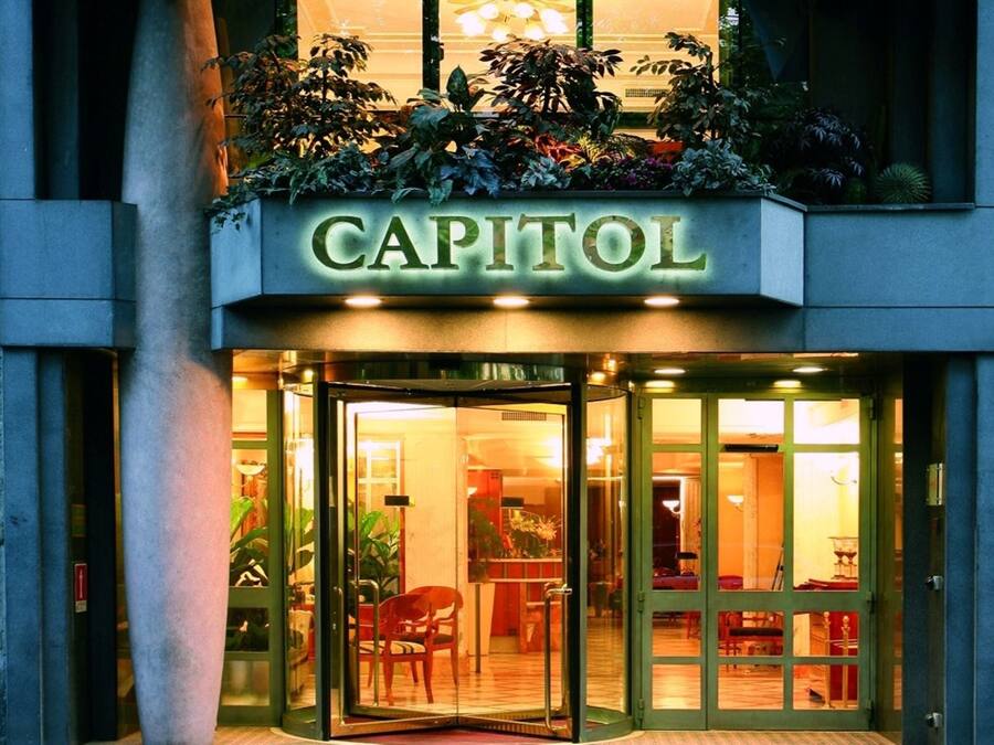Een hotelentree met een glazen deur, een bord met de tekst 'CAPITOL' en een bloembak met verschillende planten boven de ingang.