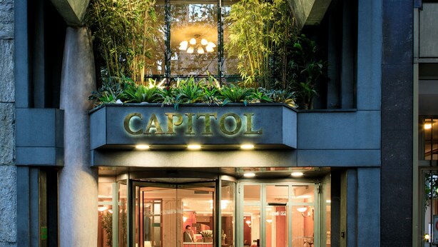 De gevel van een hotel genaamd 'Capitol', met een glazen entree, verlichte borden en een balkon met planten.