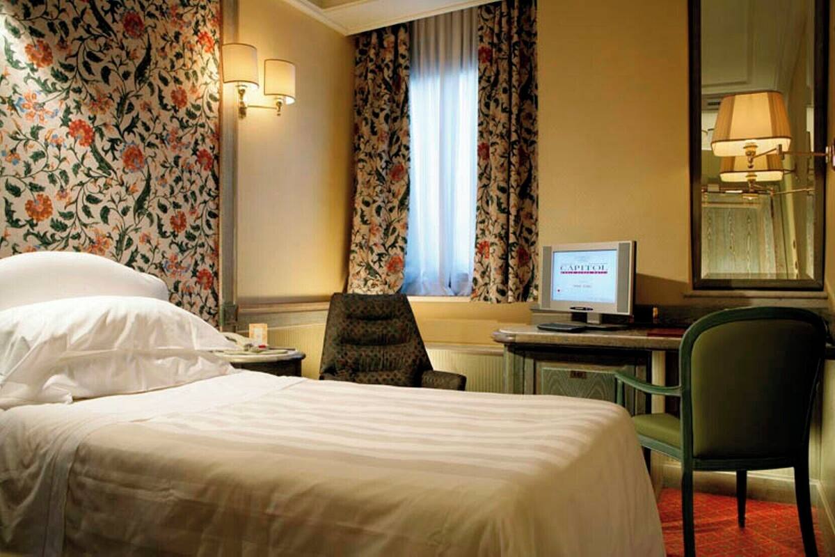 A hotel room with a bed, a desk with a computer, a chair, and floral wallpaper.