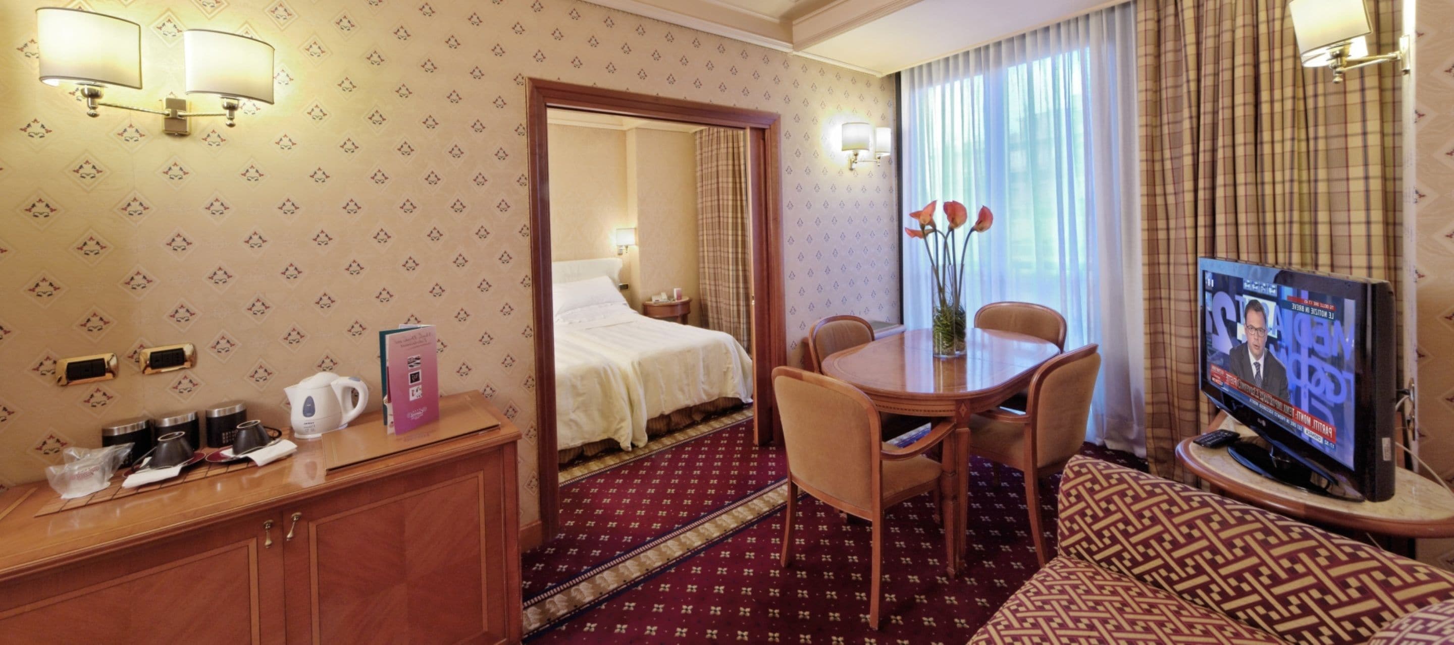 A hotel room with a bed, a dining table with chairs, a TV, and a desk with a coffee maker and books.