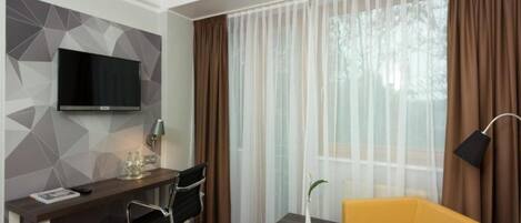 Ein Hotelzimmer mit Bett, Schreibtisch mit Stuhl, Fernseher, kleinem Tisch mit Lampe und einem Fenster mit Vorhängen.