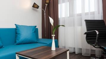 Une pièce avec un canapé bleu, un vase blanc contenant une plante, une table en bois foncé, un fauteuil de bureau noir et un lampadaire.