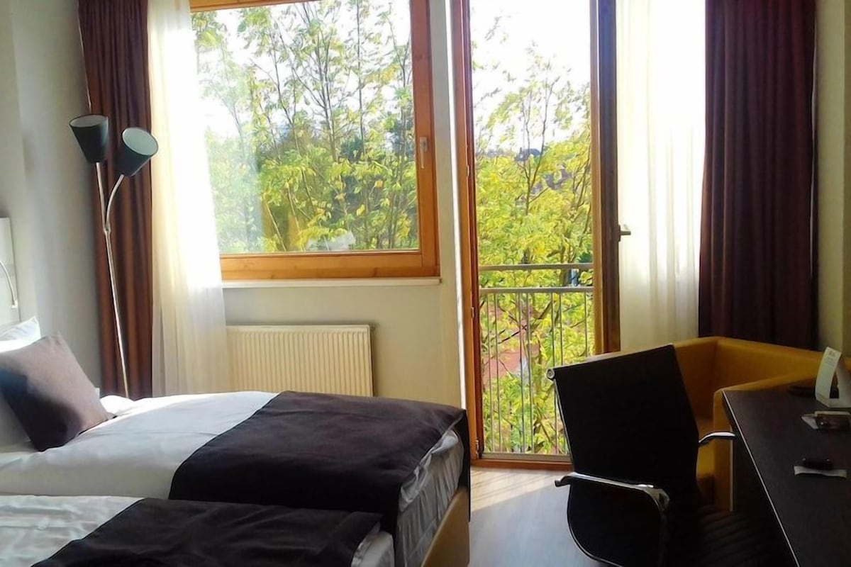 A hotel room with a bed, a desk, a chair, a window with a view of trees, and a radiator.