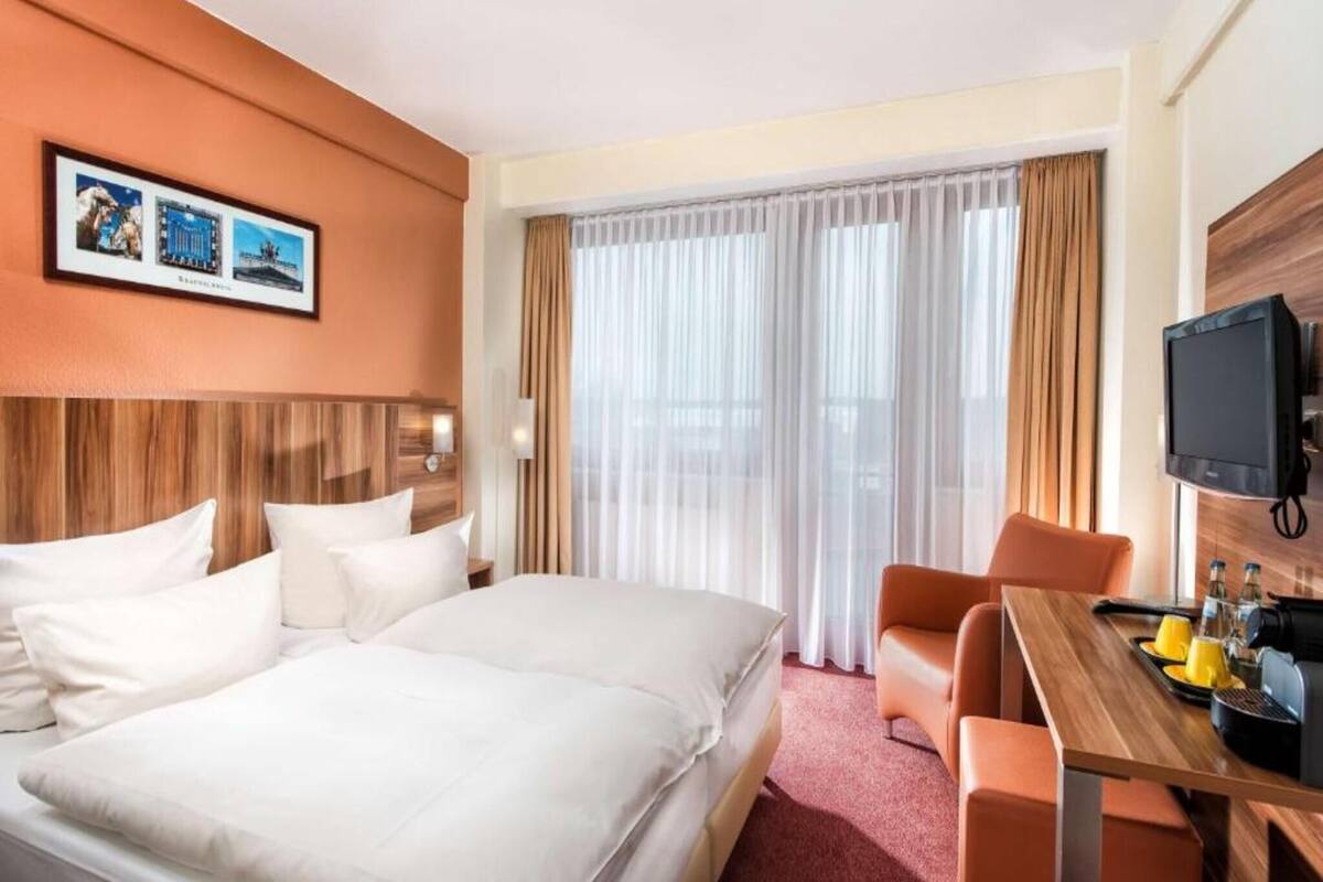 Ein Hotelzimmer mit einem großen Bett, einem Schreibtisch mit Stuhl, einem Fernseher und einem Fenster mit Vorhängen.