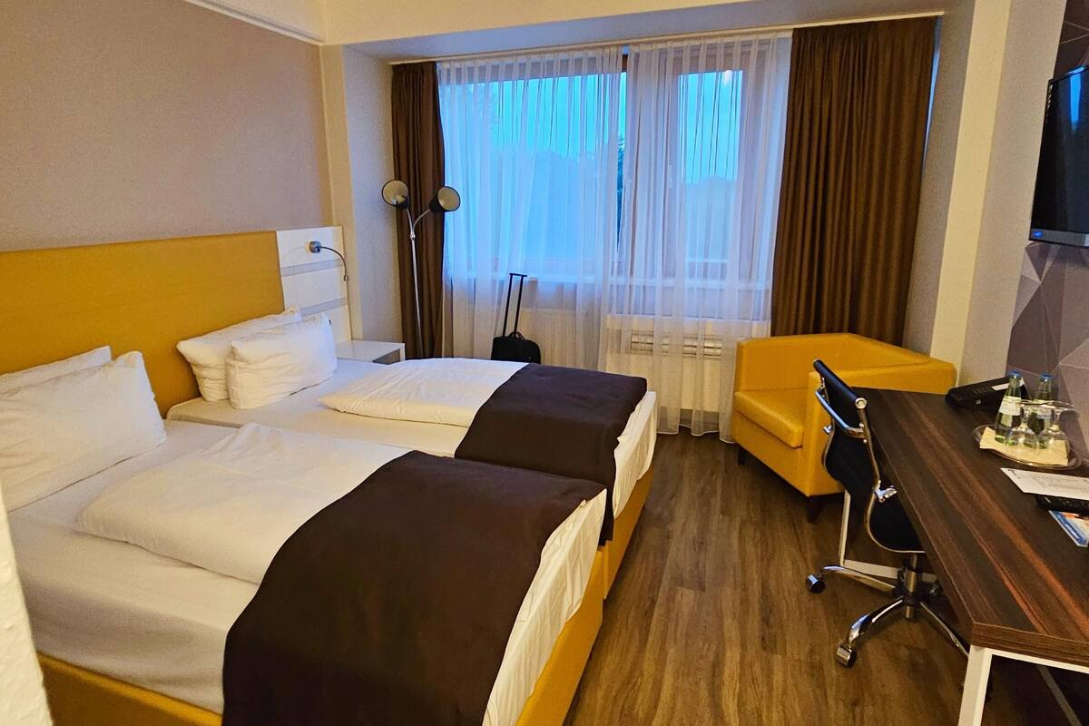 Ein Hotelzimmer mit zwei Betten, einem Schreibtisch, einem Sessel, einem Fernseher und einem Fenster mit Vorhängen.