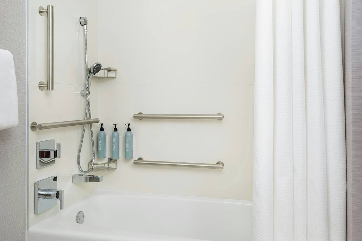 A bathroom with a bathtub, grab bars, a handheld showerhead, and two bottles of hand soap.