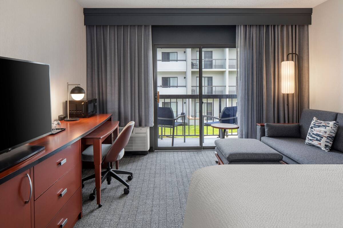 A hotel room with a bed, a desk with a TV, a chair, a sofa, and a balcony with a view of the building.
