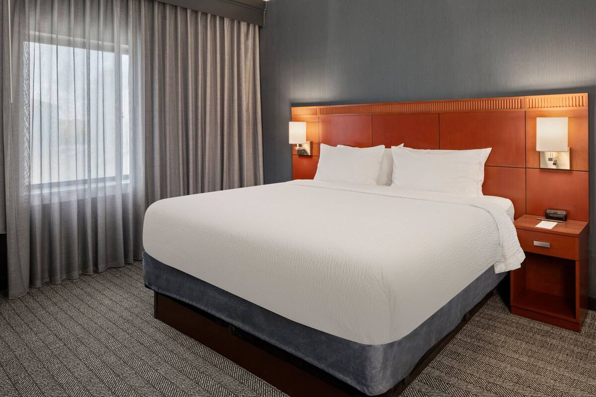 A hotel room with a large bed, a headboard with integrated lighting, a nightstand, and a window with curtains.