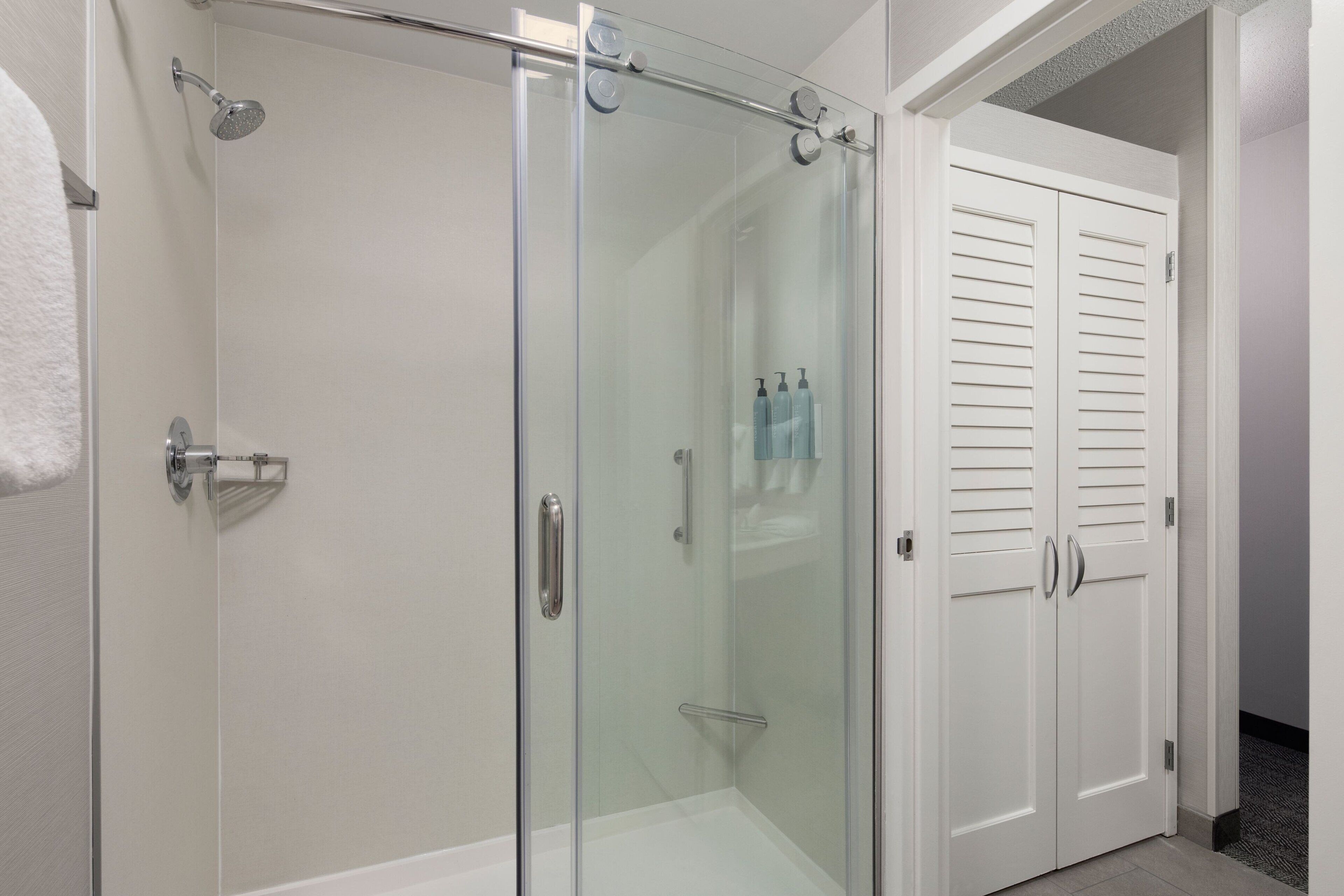 A glass-enclosed shower with a handheld showerhead, a white towel, and a white door with louvered panels.