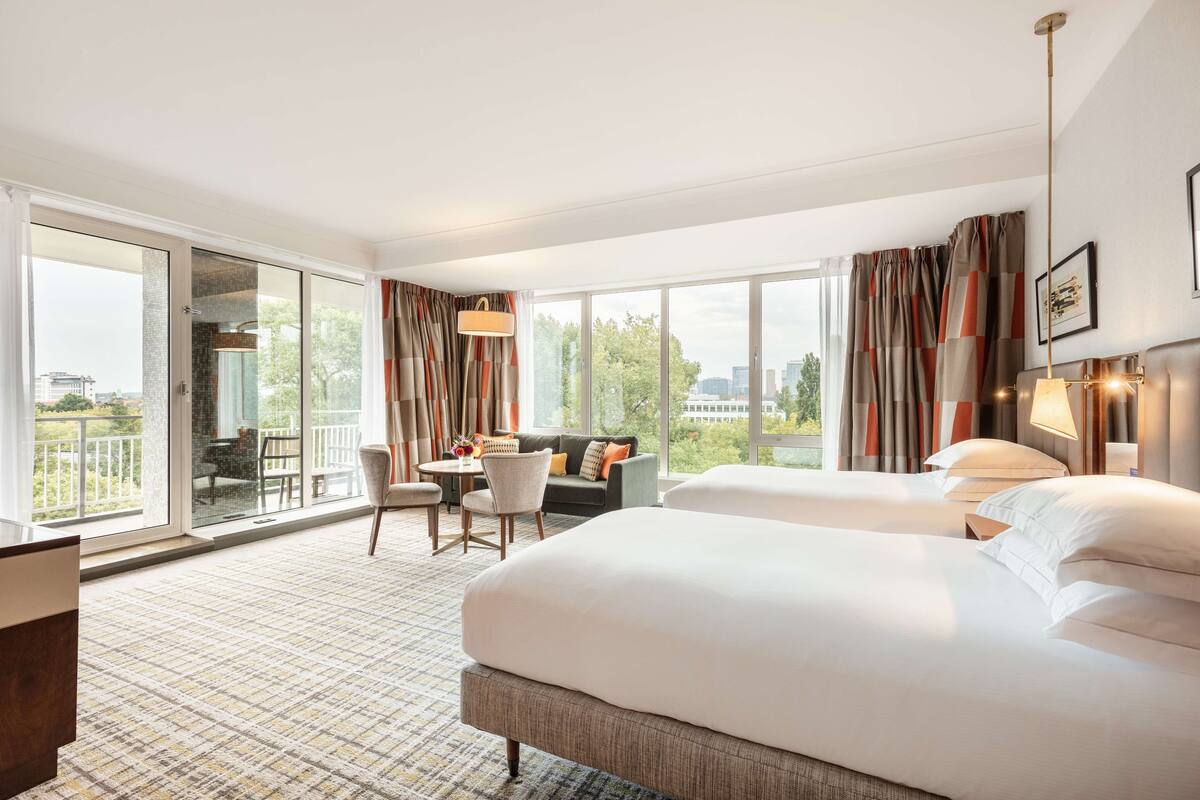 Habitación de hotel con dos camas, zona de comedor y balcón con vistas a la ciudad.