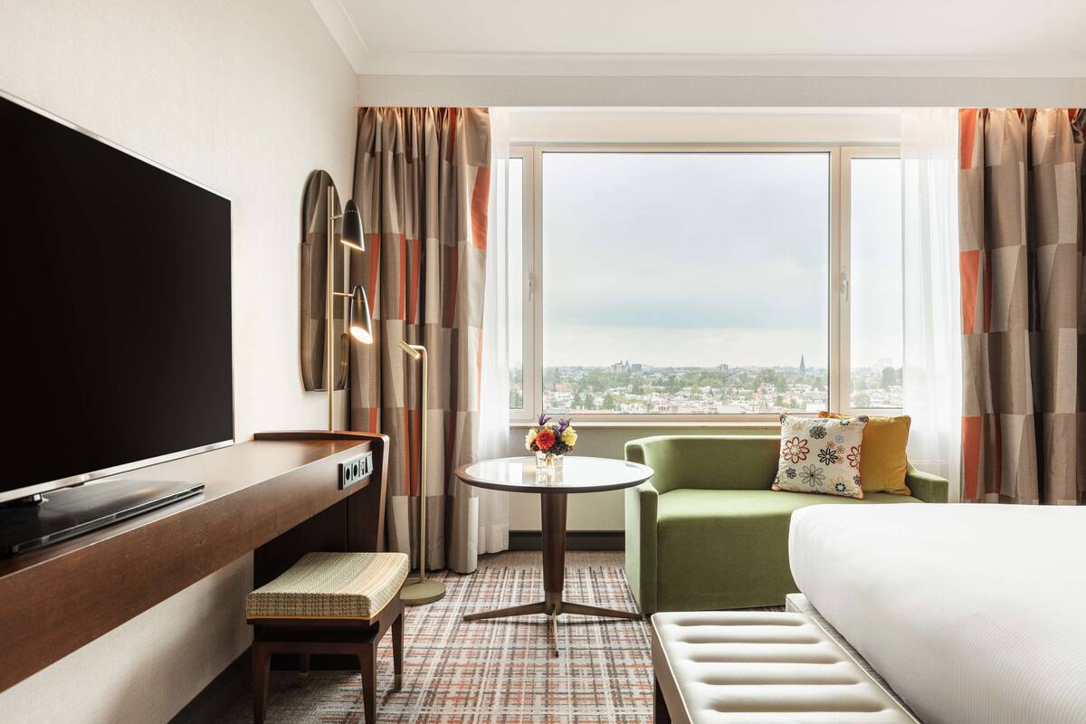 Habitación de hotel con una gran televisión de pantalla plana, un sofá verde, una cama blanca, una pequeña mesa redonda con un jarrón de flores y vistas a un paisaje urbano a través de un amplio ventanal.