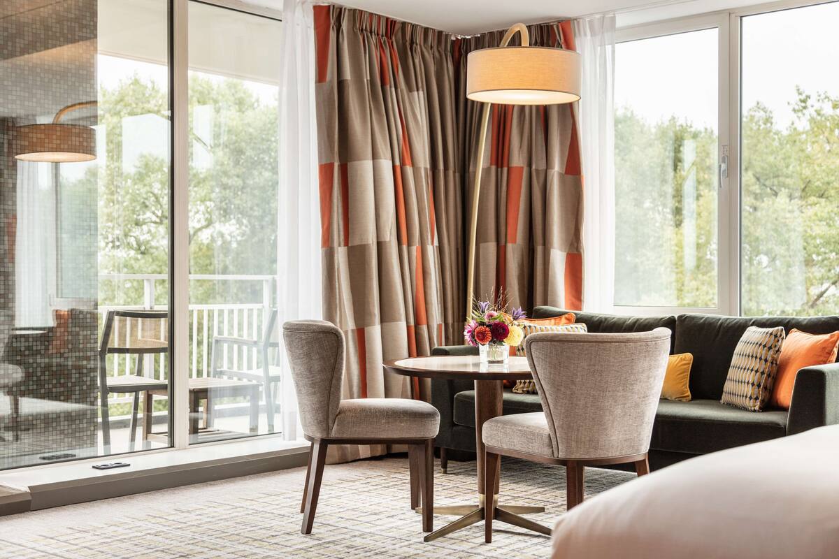 Una habitación de hotel moderna con un ventanal grande, una zona de comedor con mesa y sillas, y un sofá con cojines.