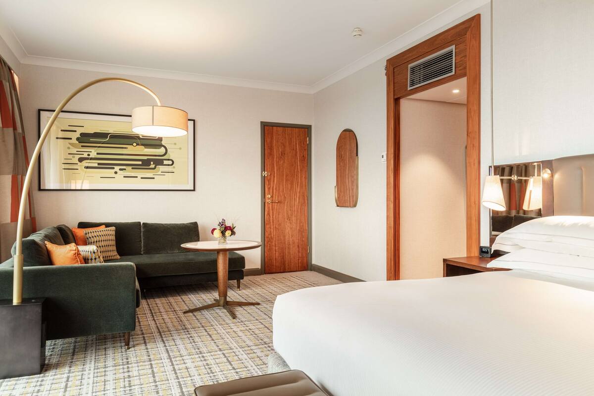 Habitación de hotel con una cama grande, un sofá, una mesita con un jarrón de flores y un cuadro en la pared.