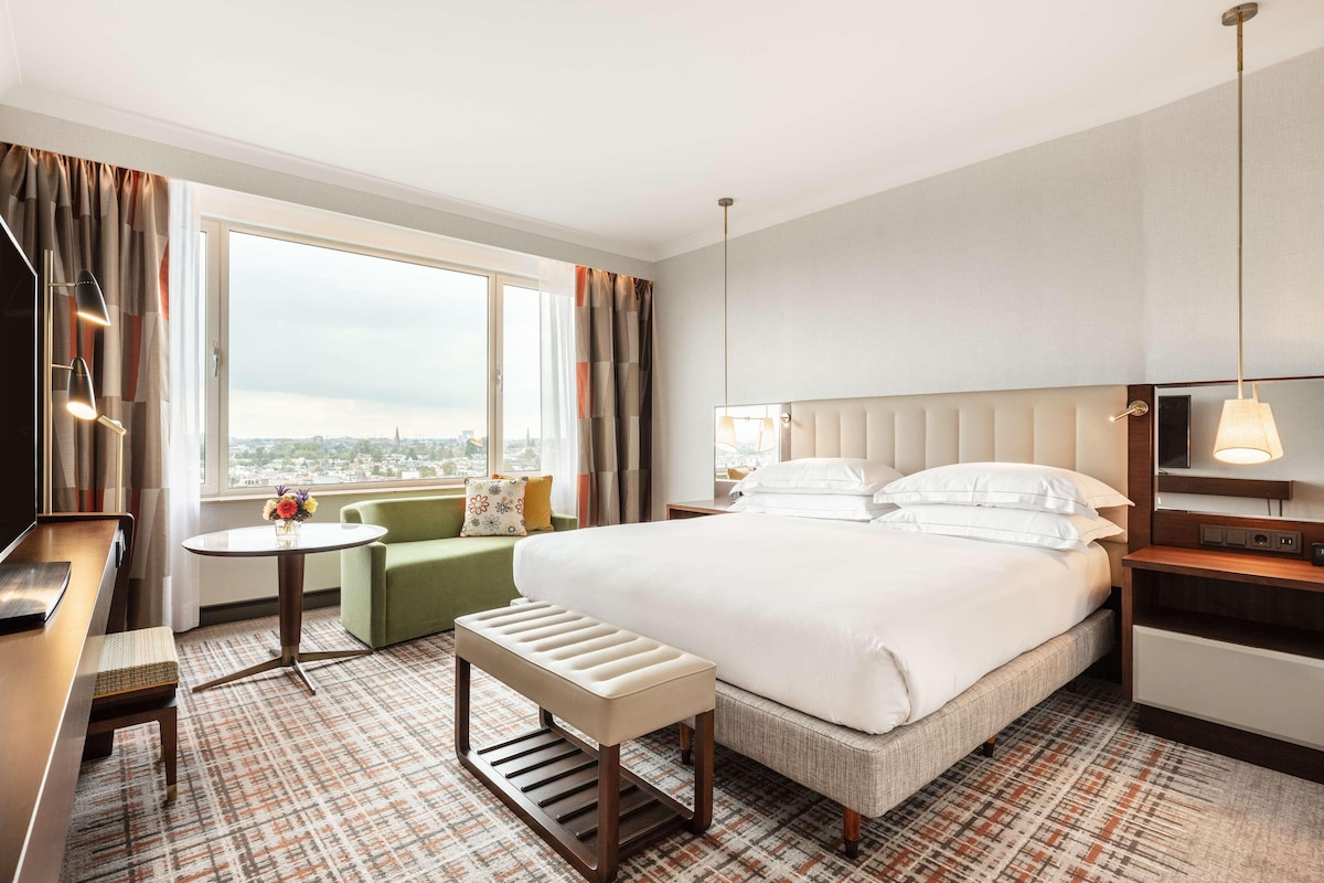 Habitación de hotel con una cama grande, un escritorio, una silla, una mesita con un jarrón y vistas a la ciudad.