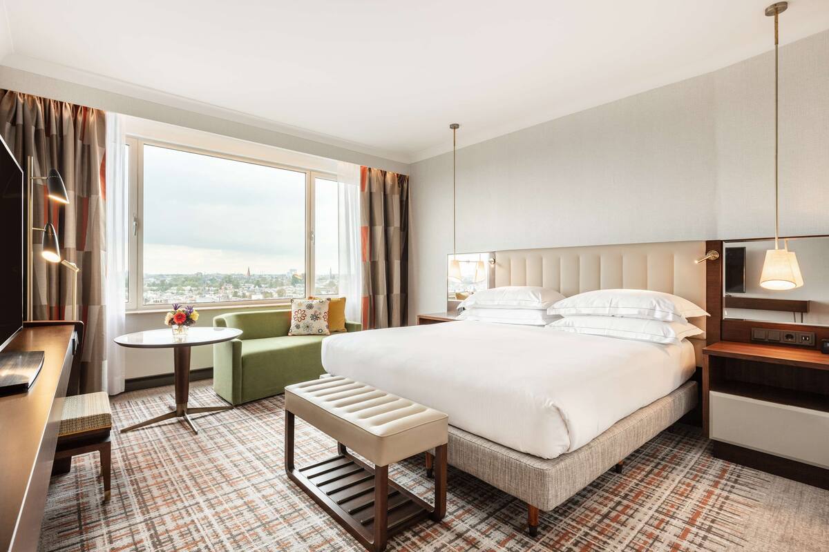 Habitación de hotel con una cama grande, un escritorio, una silla, una mesita con un jarrón y vistas a la ciudad.