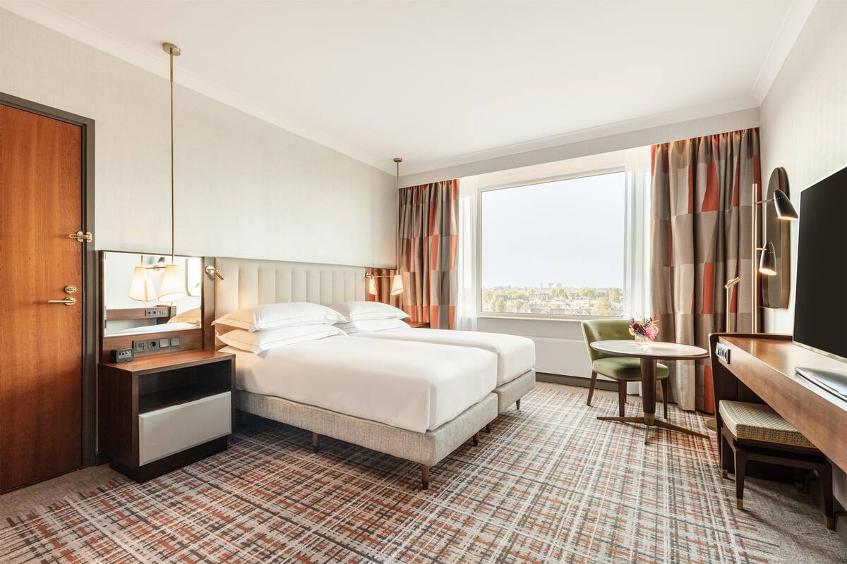Habitación de hotel con cama, mesita de noche, lámpara, televisor y vista a la ciudad.