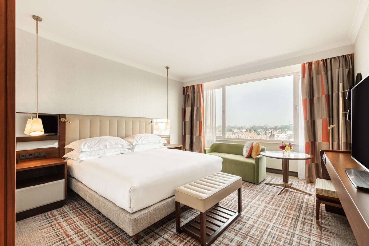 Habitación de hotel con una cama grande, un sillón verde, una mesita redonda y un televisor.