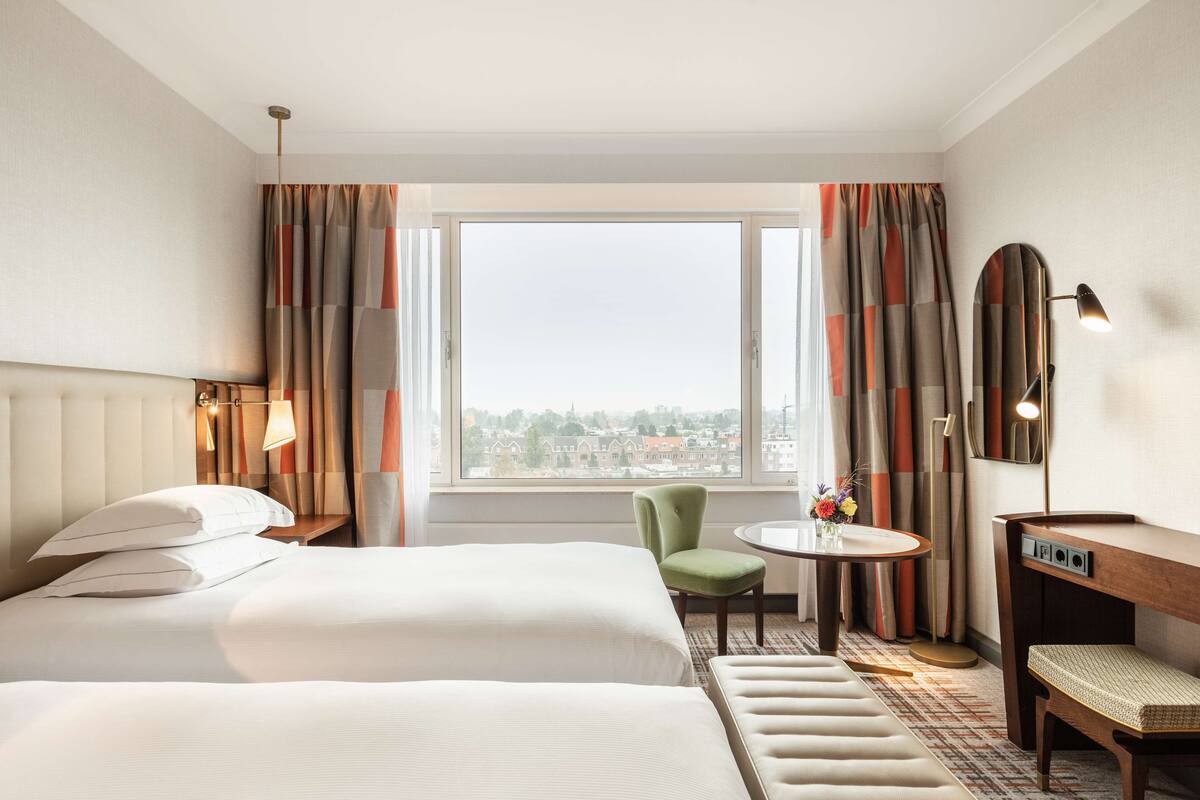 Habitación de hotel con dos camas, una mesita con un florero, una silla, un escritorio y vistas de la ciudad a través de amplias ventanas.