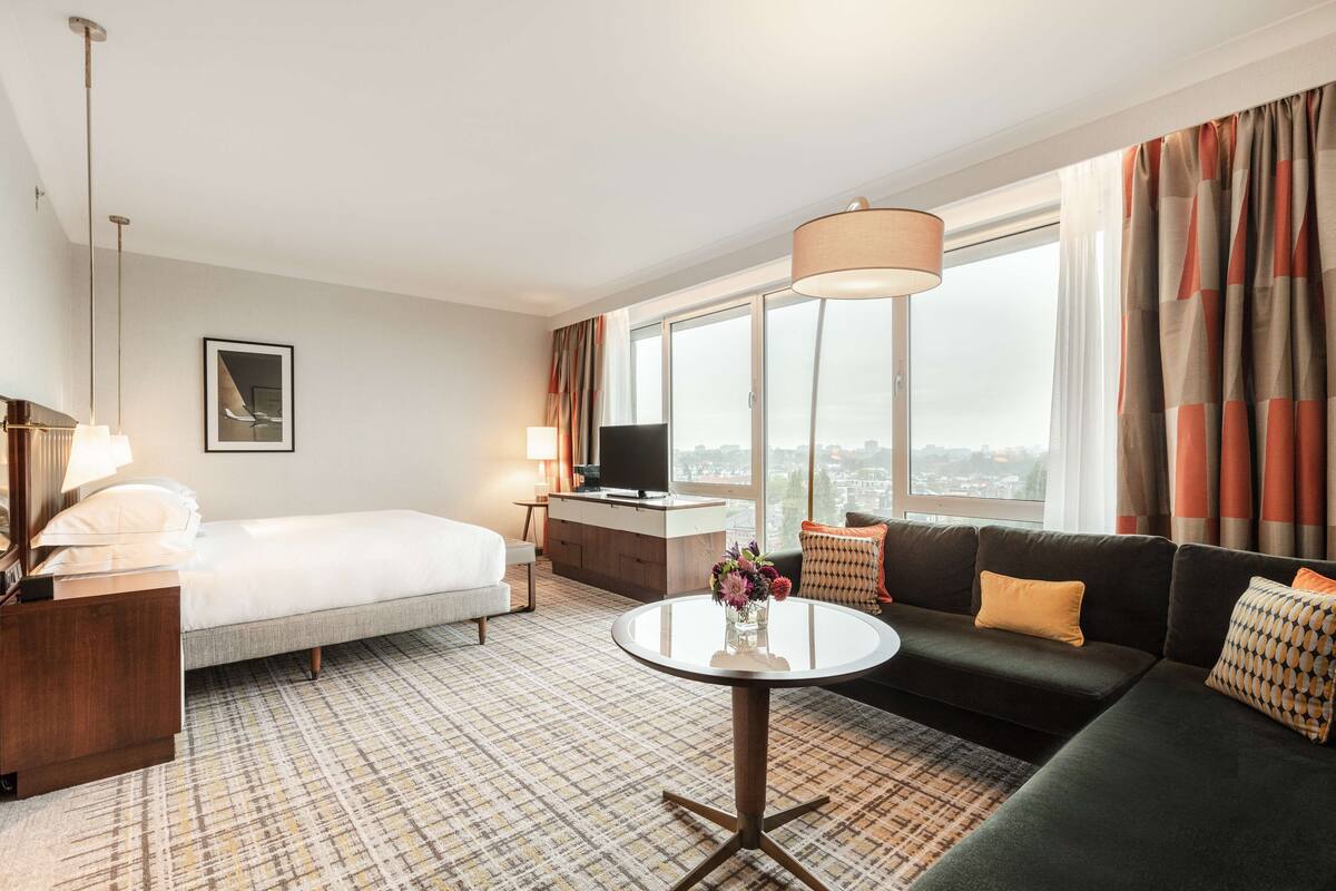 Habitación de hotel con cama, sofá, una mesa redonda, televisor y un ventanal con cortinas.