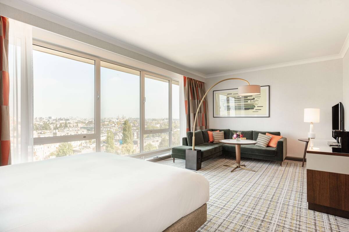 Una habitación de hotel con una cama grande, un sofá, una mesita y un escritorio con una lámpara. Una ventana grande ofrece vistas a la ciudad.