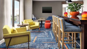 A modern lounge area with yellow and blue patterned sofas, a bar with wooden chairs, a flat-screen TV, and a large potted plant. - Hampton Inn Laredo (Laredo)