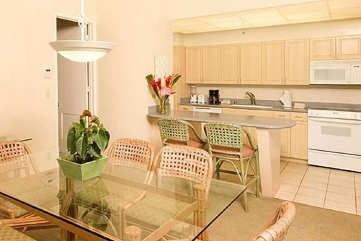A dining area with a glass table and wicker chairs, a kitchen with wooden cabinets and white appliances, and a small island with a sink.