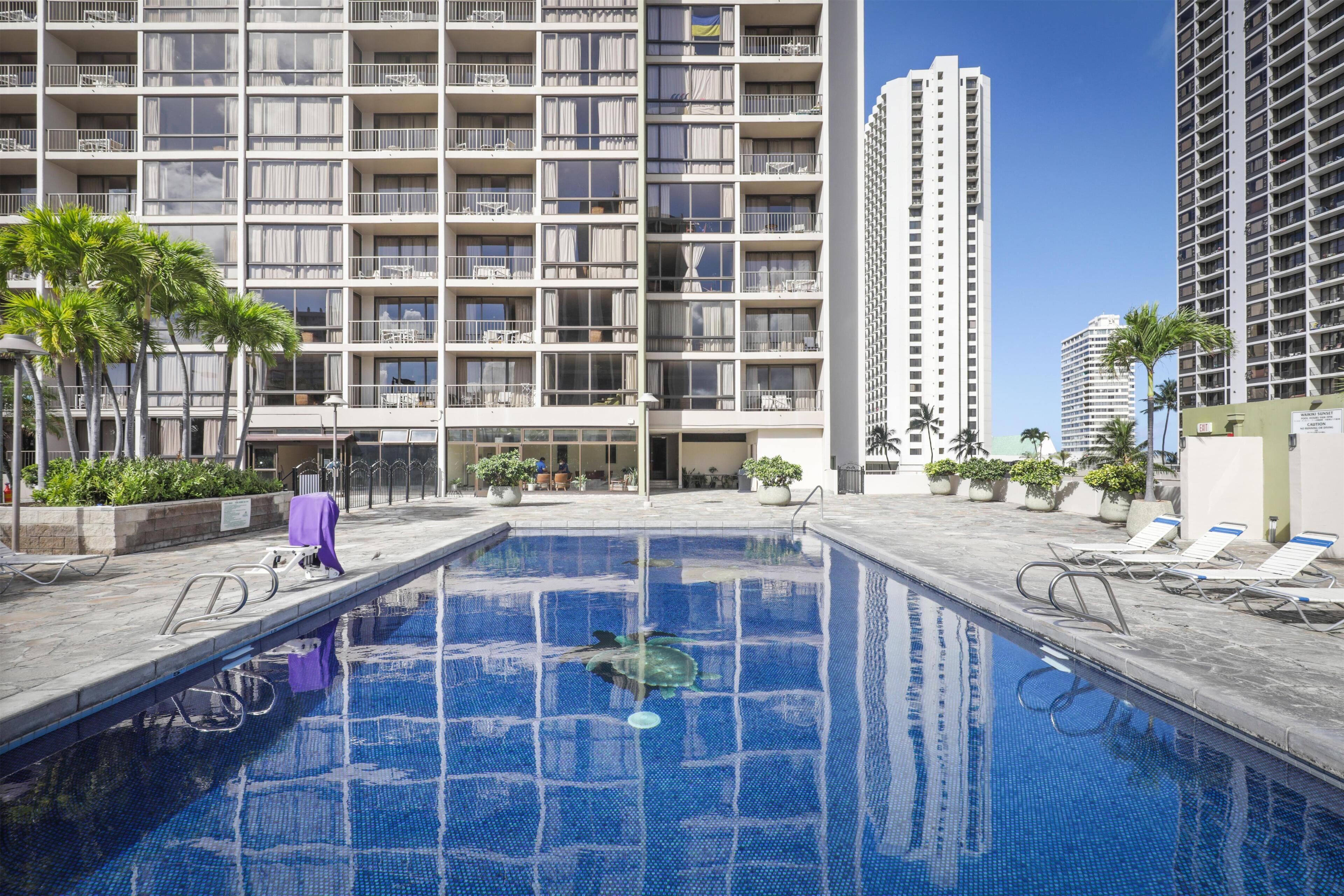 A hotel pool area with a clear view of the pool, lounge chairs, and a high-rise building in the background.