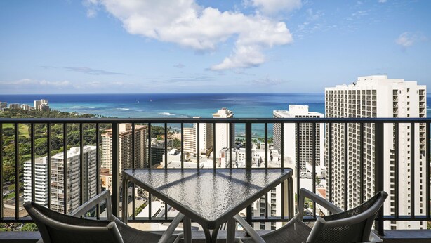 A balcony with a table and chairs overlooking a cityscape and ocean. - Aston Waikiki Sunset (Honolulu)