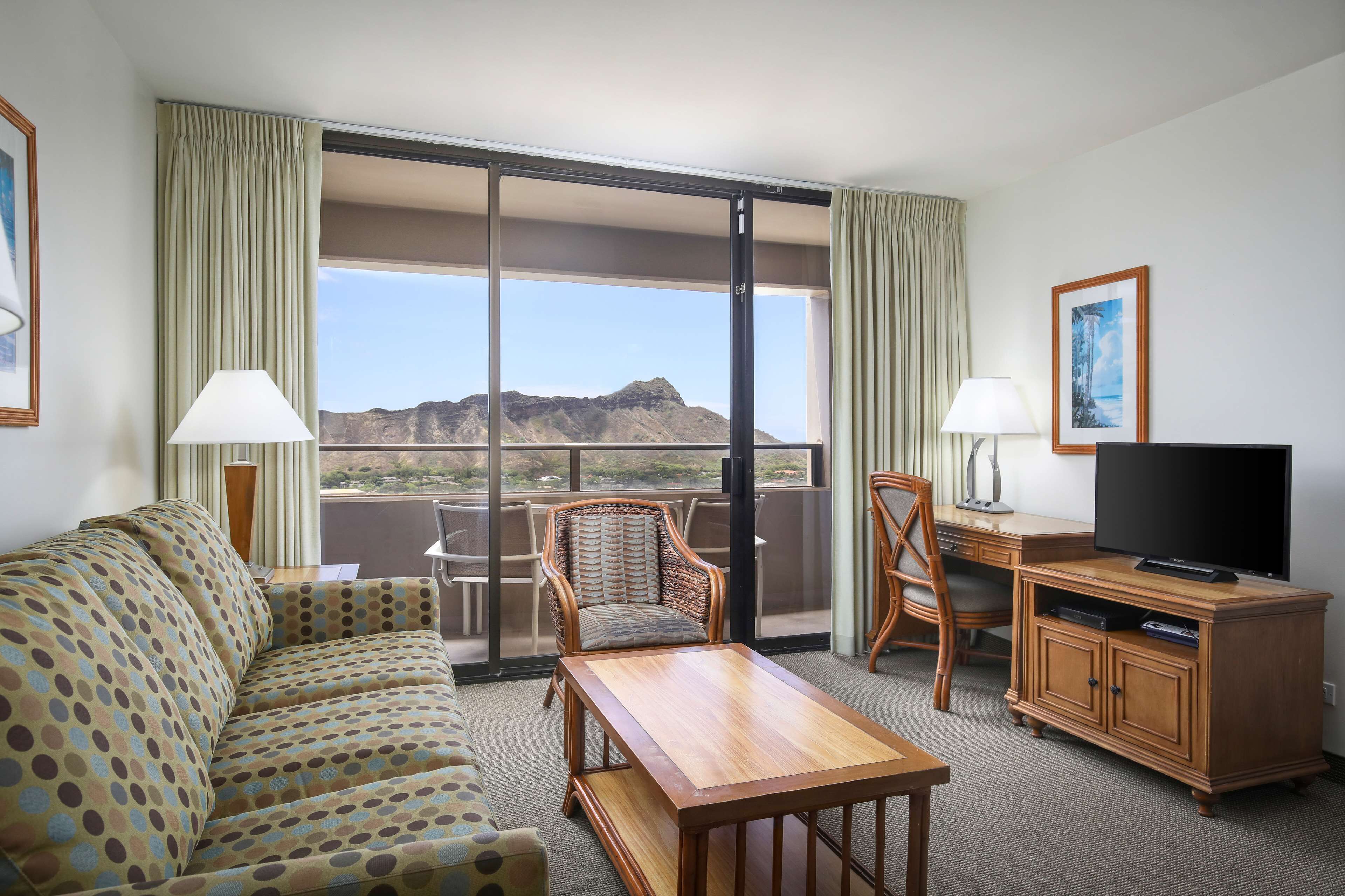 A hotel room with a sofa, a wooden table, a TV, and a desk. There is a large window with a view of mountains.