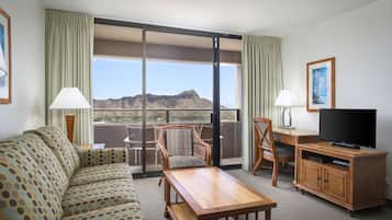 A hotel room with a sofa, a wooden table, a TV, and a desk. There is a large window with a view of mountains.