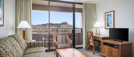 Quarto de hotel com sofá, mesa de madeira, televisão e escrivaninha. Possui uma janela grande com vista para montanhas.