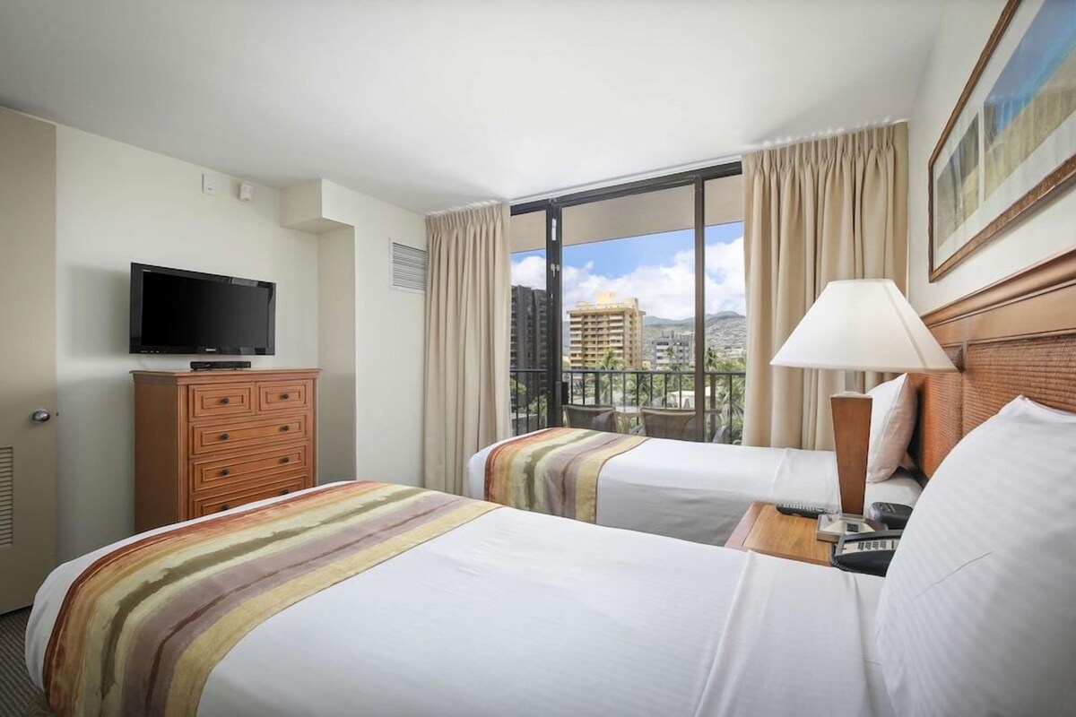 A hotel room with two beds, a television, a dresser, and a large window with a view of buildings.