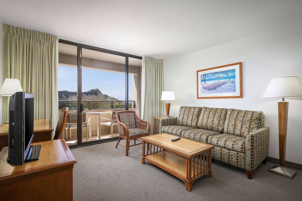 A hotel room with a sofa, armchair, TV, and a balcony with a view of the mountains.