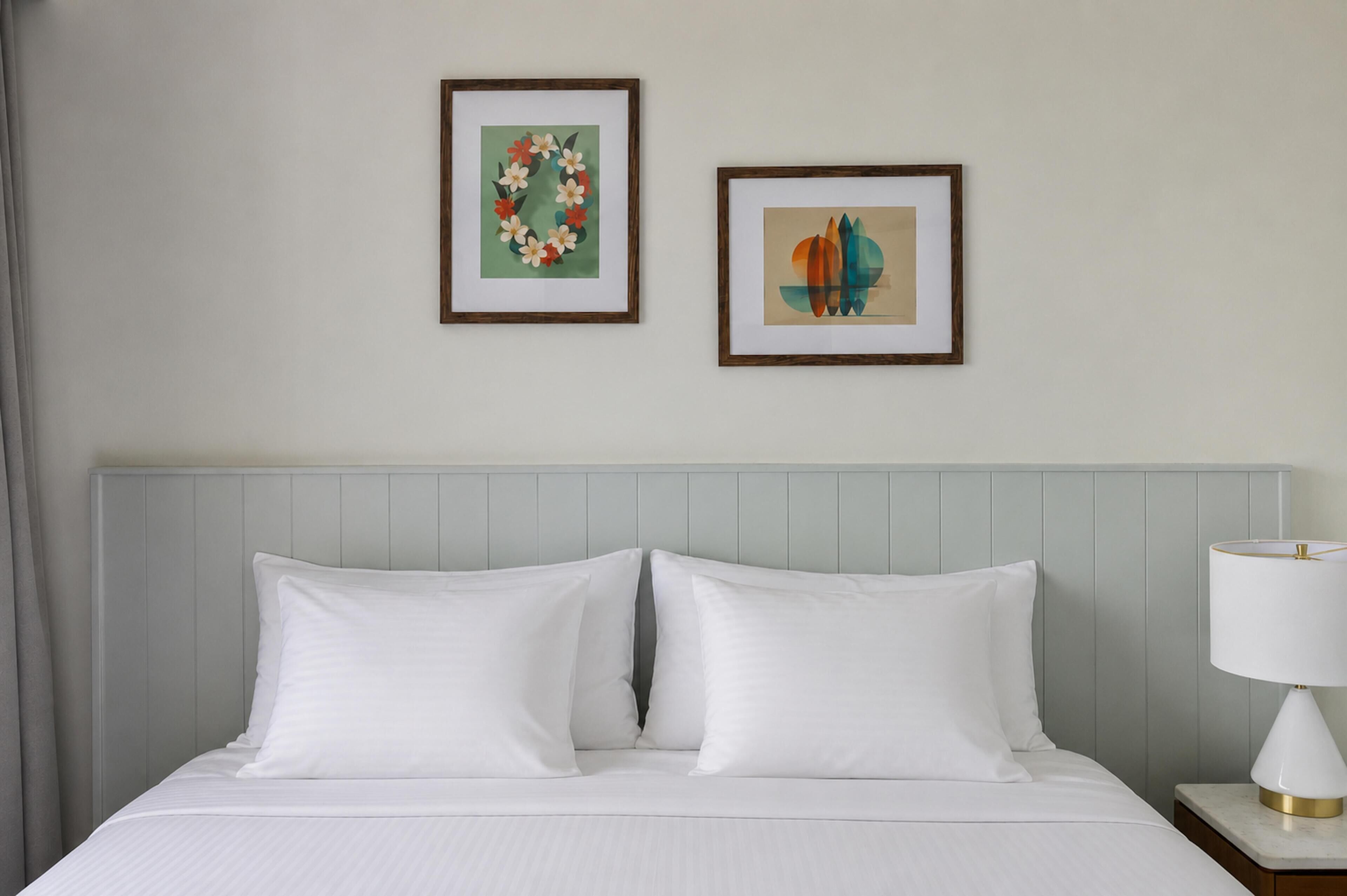 Een hotelkamer met een bed, twee kussens, een nachtkastje met een lamp en twee kunstwerken in lijsten aan de muur.