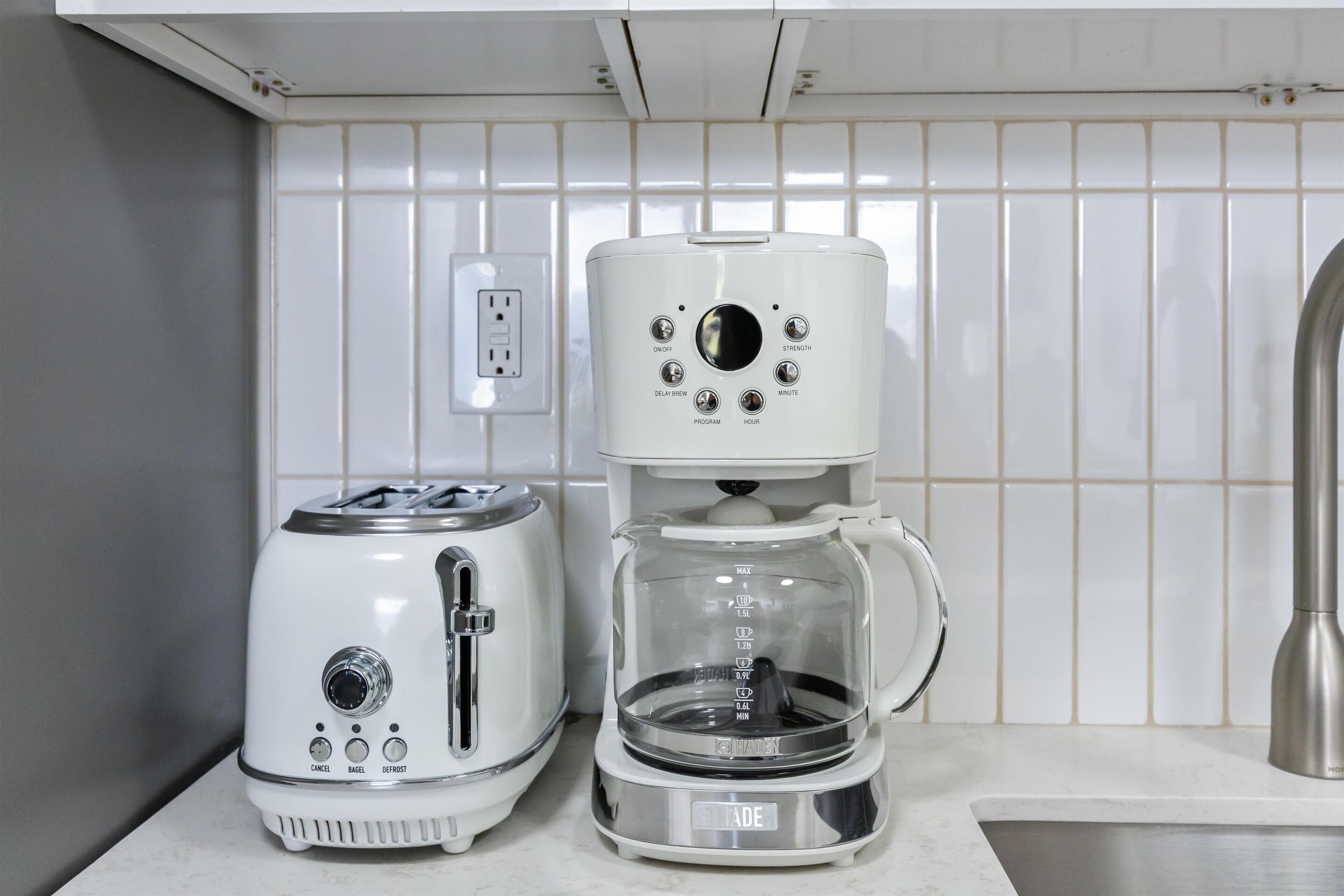 A kitchen with a coffee maker and toaster on the counter, a power outlet on the wall, and a stainless steel sink.