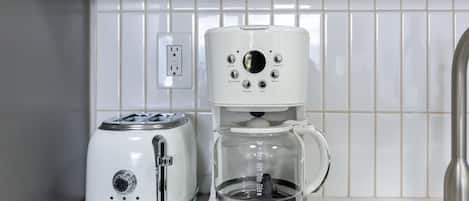 A kitchen with a coffee maker and toaster on the counter, a power outlet on the wall, and a stainless steel sink.