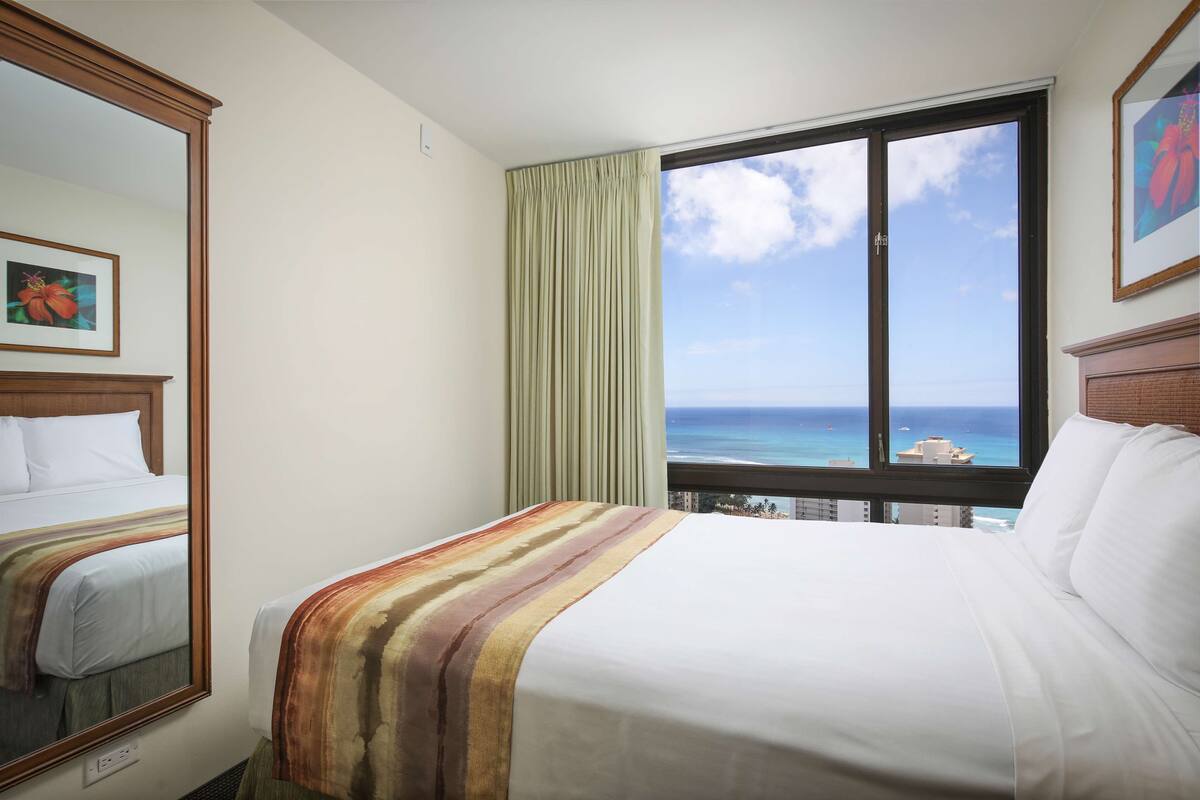 A hotel room with a bed, a window with a view of the ocean, a mirror, and a framed picture on the wall.