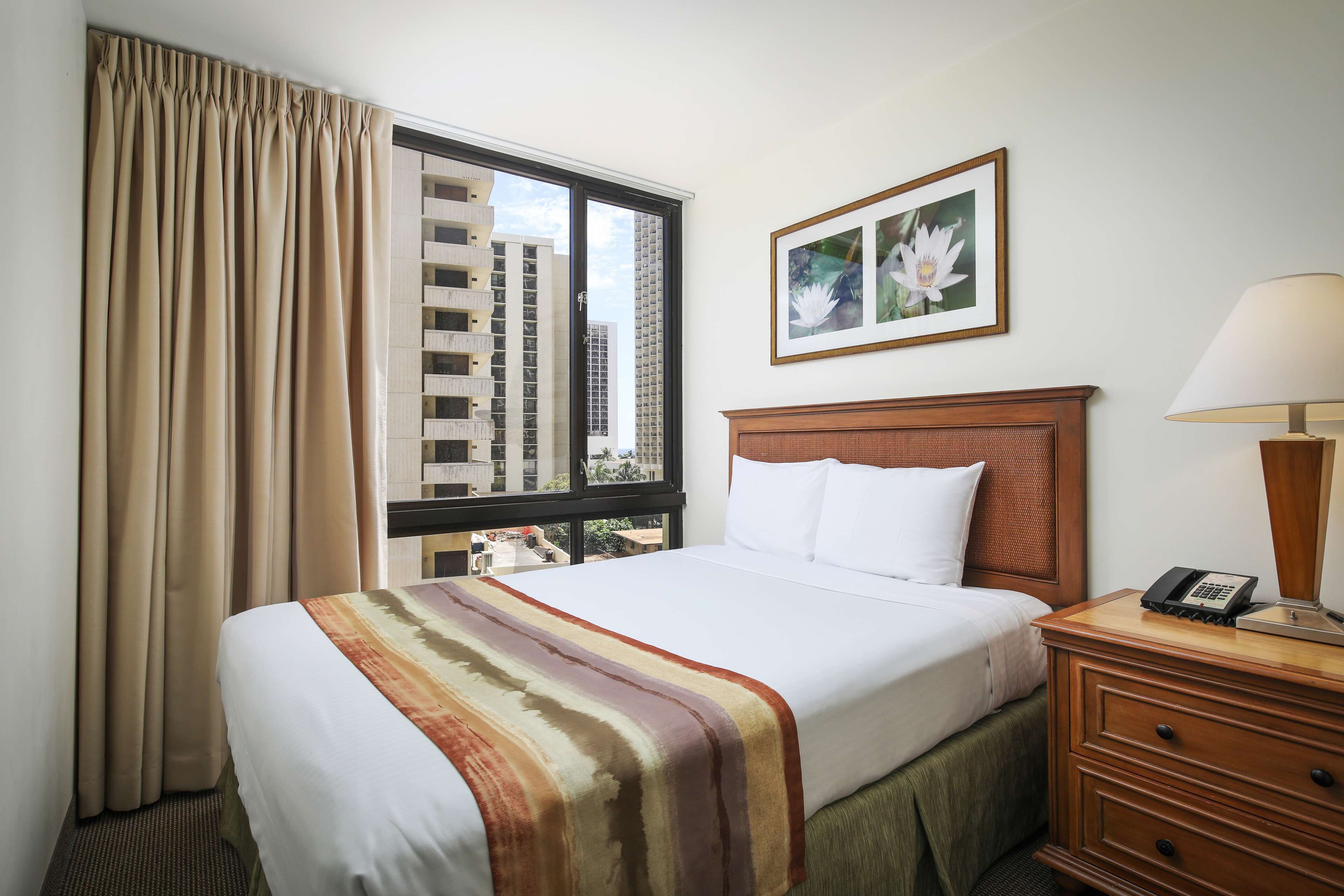 Habitación de hotel con una cama grande, una mesita de noche de madera con una lámpara, un cuadro enmarcado en la pared y una ventana con cortinas.