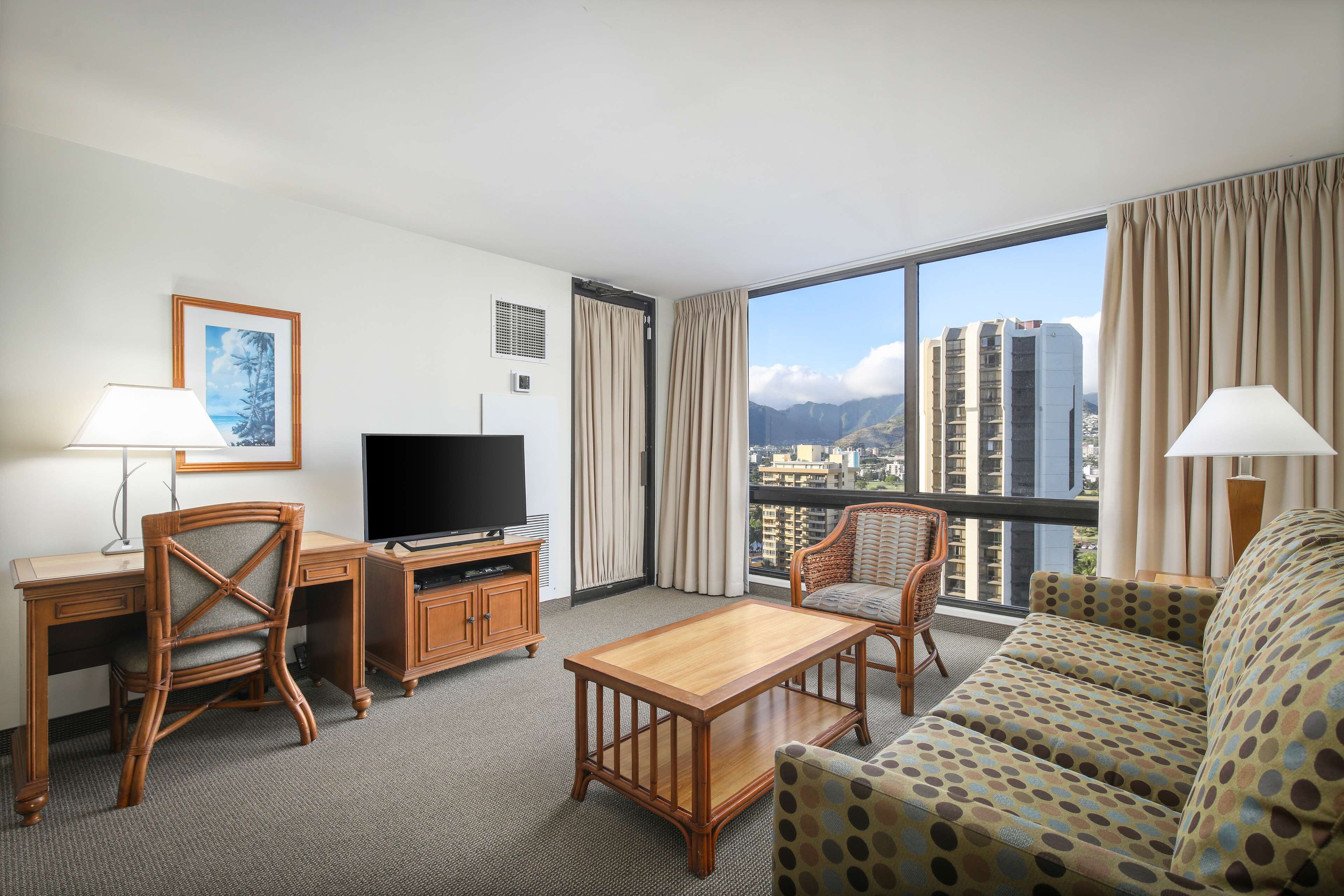 A hotel room with a large window, a television, a desk with a lamp, a sofa, and a chair.