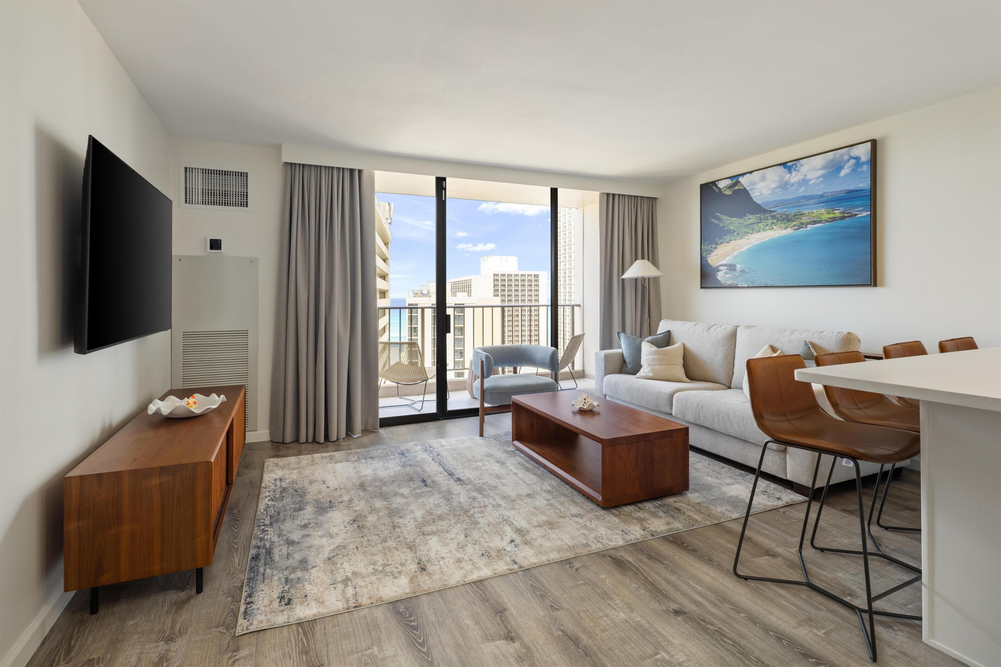 A modern hotel room with a large window, a flat-screen TV, a sofa, a coffee table, and a dining area with chairs.