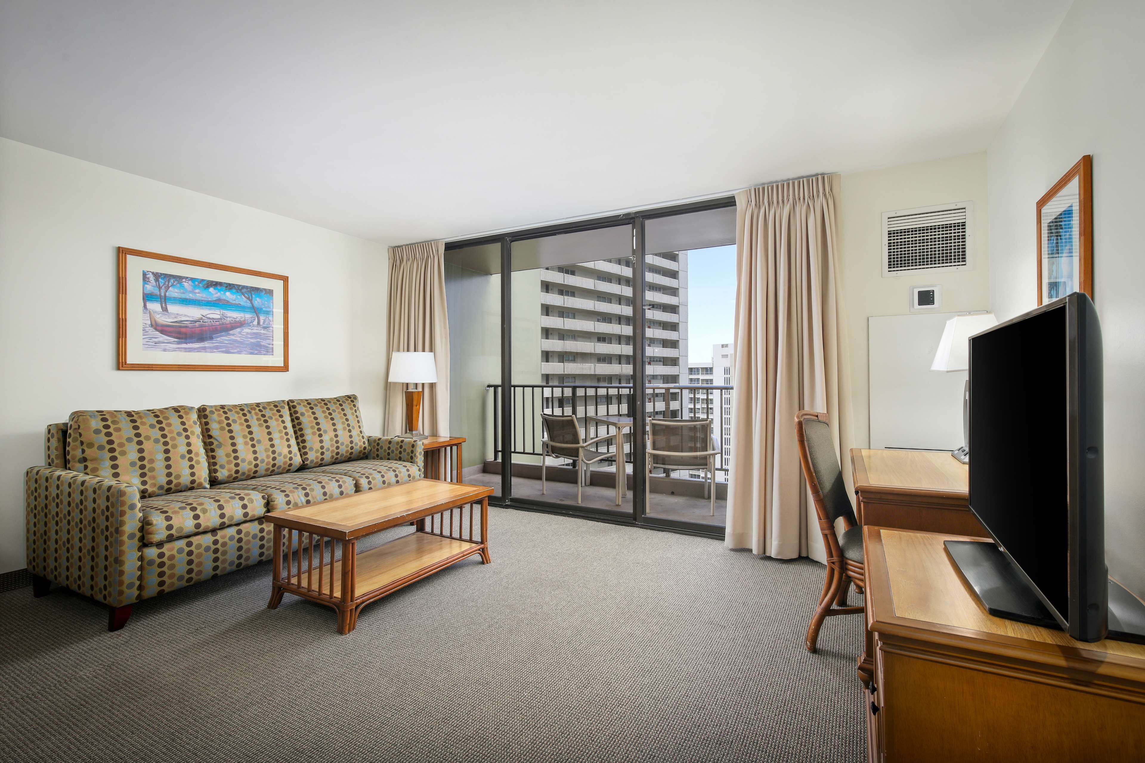 A hotel room with a sofa, a wooden coffee table, a TV, and a balcony with a view of a building.
