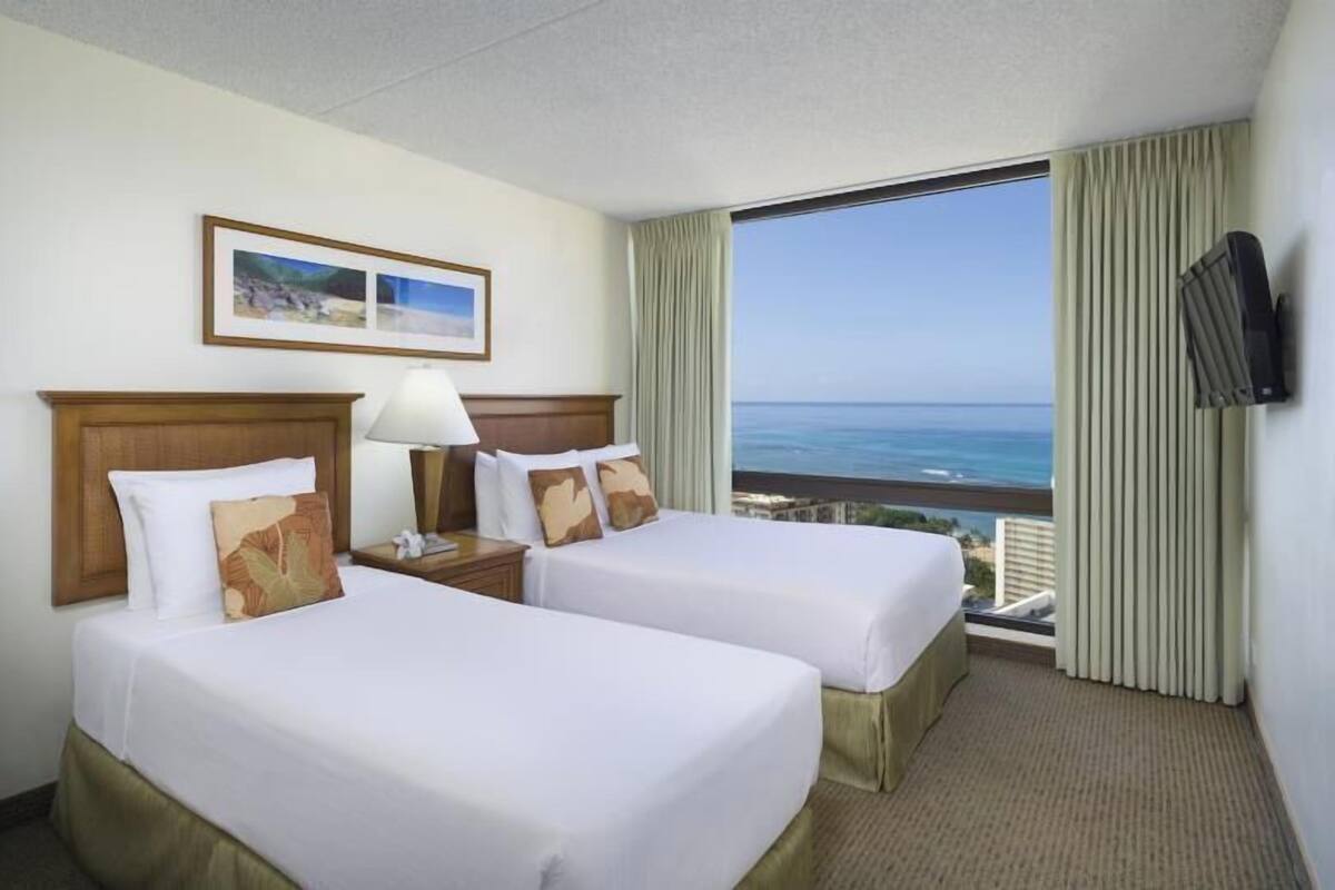 A hotel room with two beds, a nightstand, a lamp, a TV, and a large window with a view of the ocean.