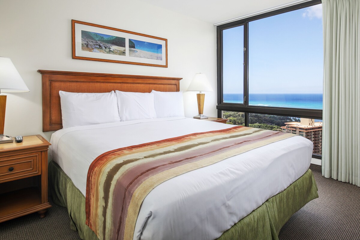 Quarto de hotel com uma cama grande, uma mesinha de cabeceira, uma luminária, um quadro na parede e vista para o oceano pela janela.