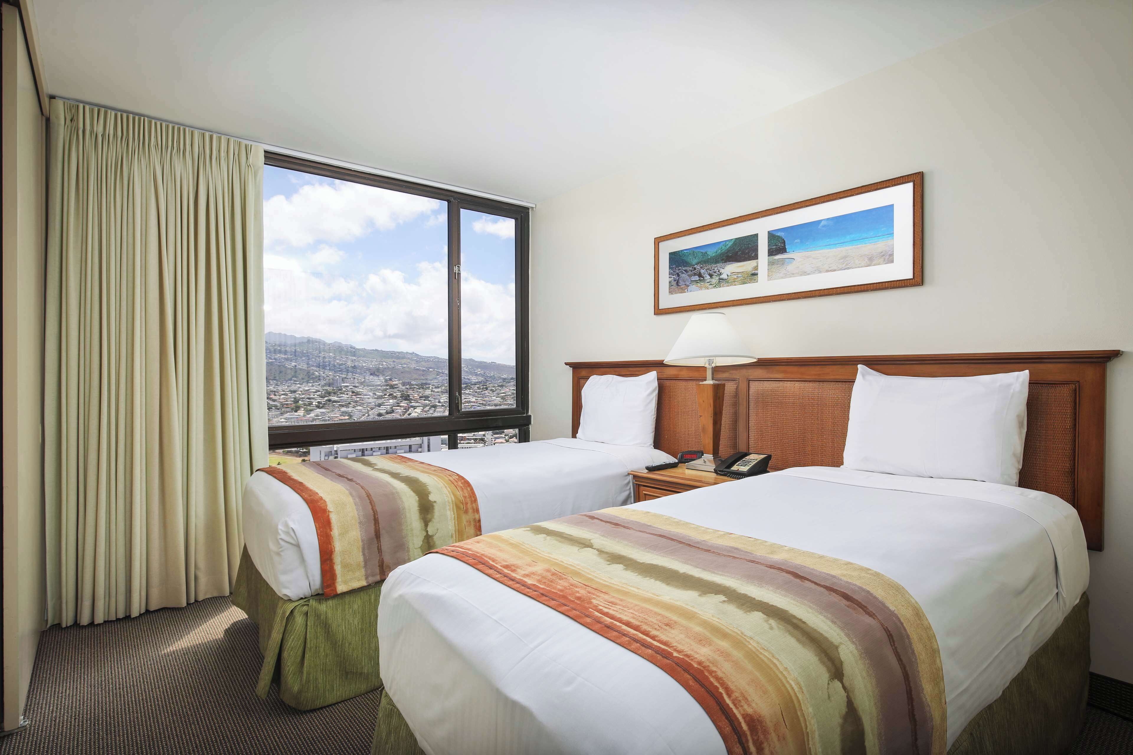 Ein Hotelzimmer mit zwei Betten, einem großen Fenster mit Ausblick, einem Nachttisch mit Lampe und einem gerahmten Bild an der Wand.