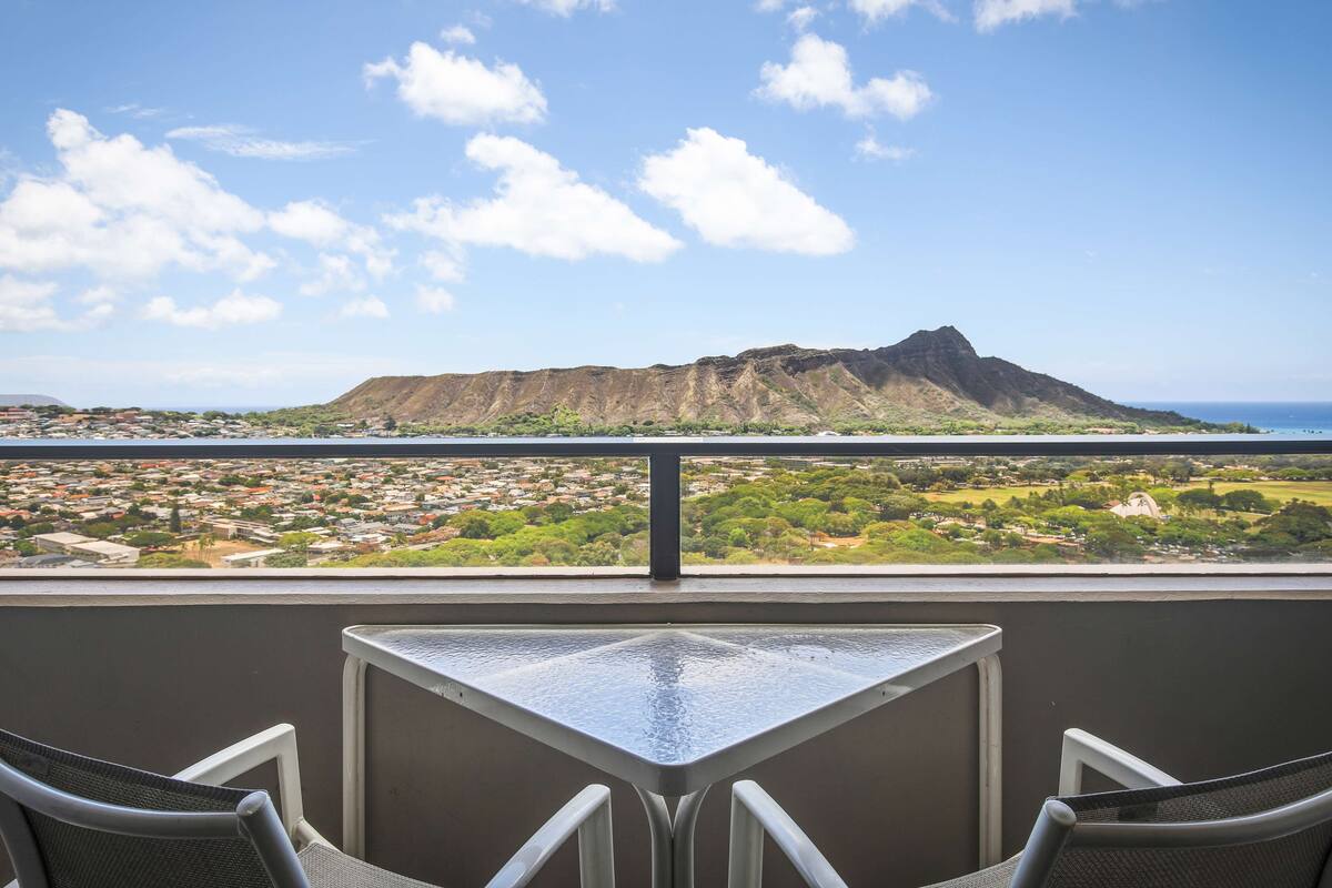 A balcony with a table and chairs, overlooking a residential area and a mountain.