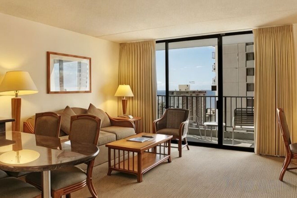 A hotel room with a sofa, armchairs, a small table, and a balcony with a view.