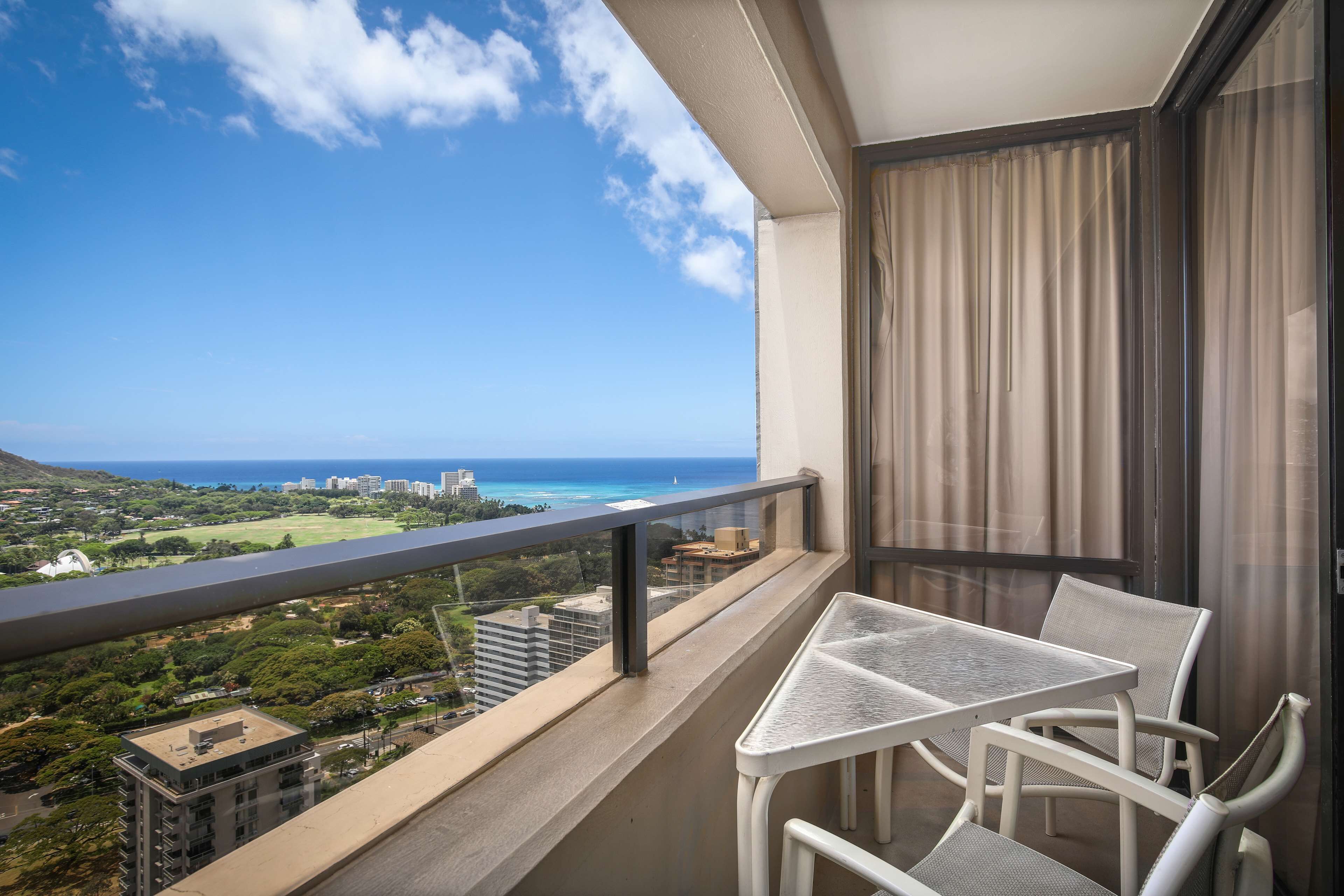 A balcony with a table and chairs, overlooking a coastal view with buildings and greenery.