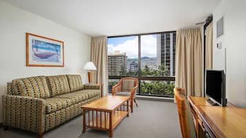 Habitación de hotel con sofá, una mesa de centro de madera, una silla de mimbre, un televisor y un ventanal con vista a la ciudad.